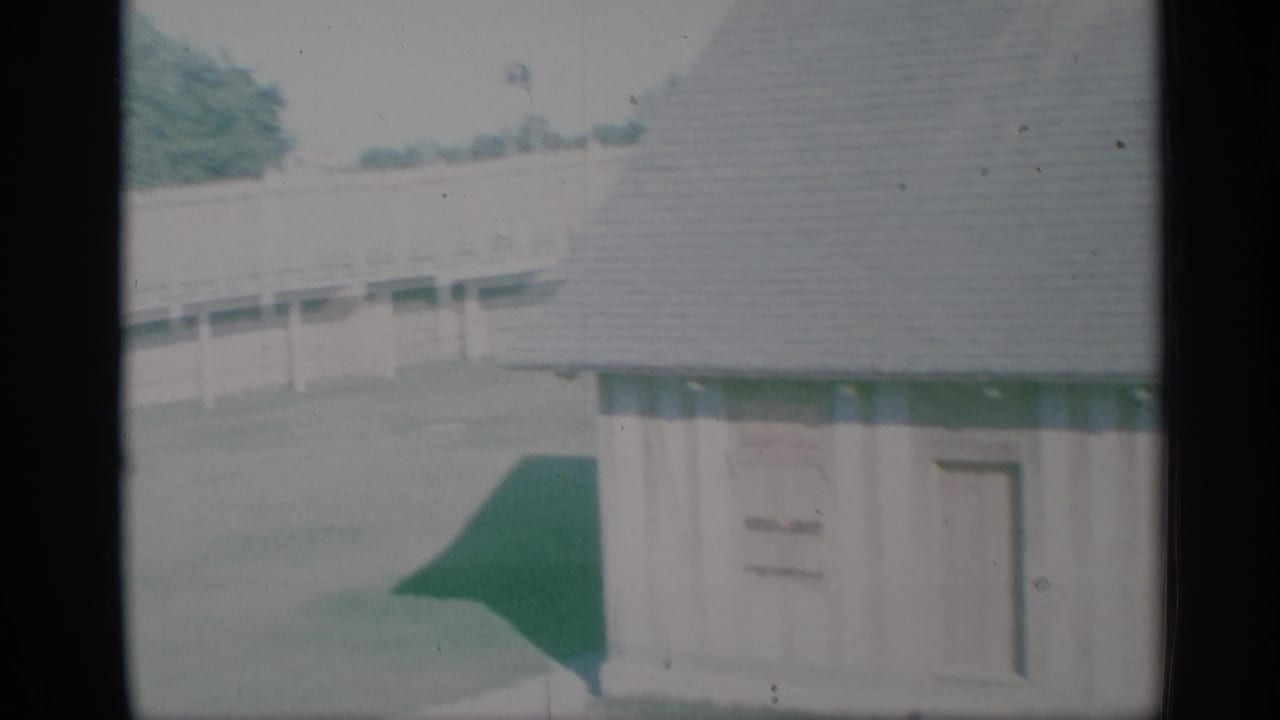 1959 New York home-movie view of the house corner, green-trimmed entry area, porch rail, lawn, and shingled roof.
