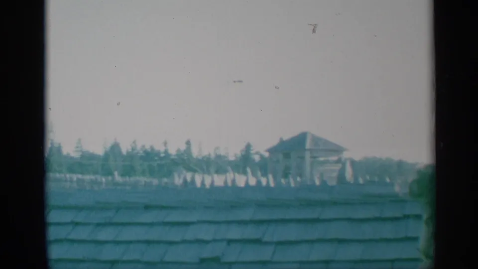 1959 New York 8mm view across a shingled rooftop toward pale fort walls, evergreen trees, and a small tower-like structure.