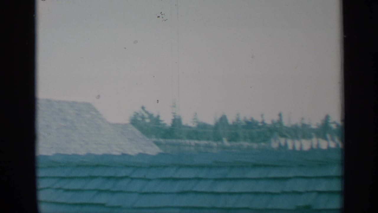 1959 New York 8mm frame with a broad shingled roof foreground, pale fort wall, distant trees, and a partial roof edge during the pan.
