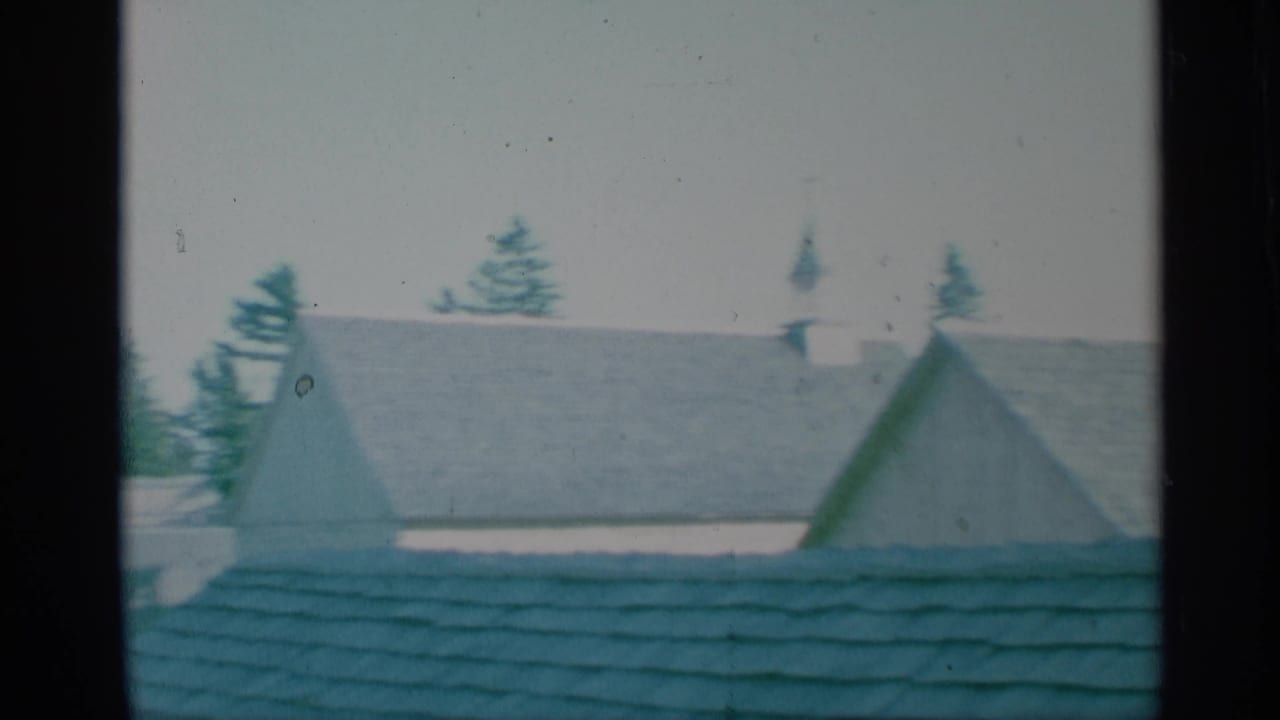 Rooftops, evergreen trees, and a distant church steeple seen through dark film-frame edges.