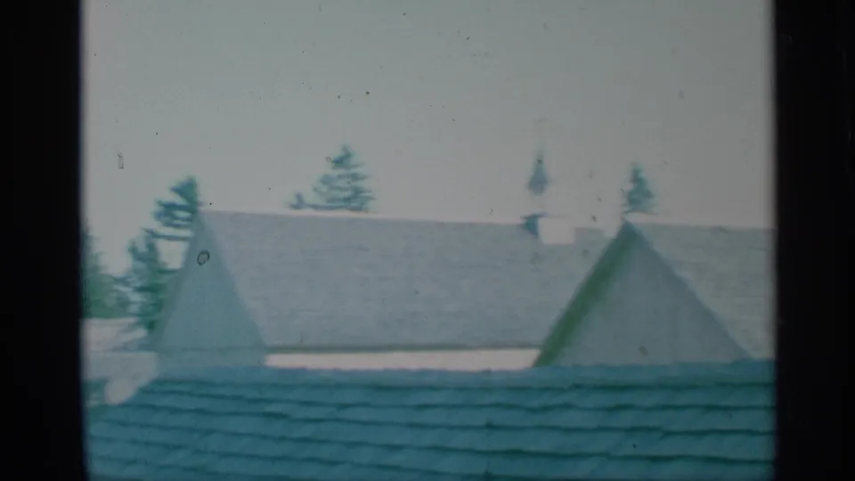 1959 New York 8mm view of shingled rooftops, evergreen trees, and a church steeple in a hazy suburban exterior.