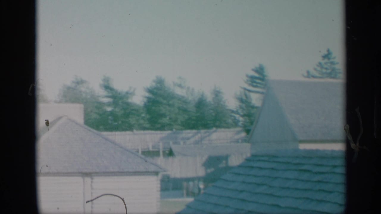 1959 New York home-movie frame with pale buildings, rooftops, evergreen trees, and a church steeple behind the houses.