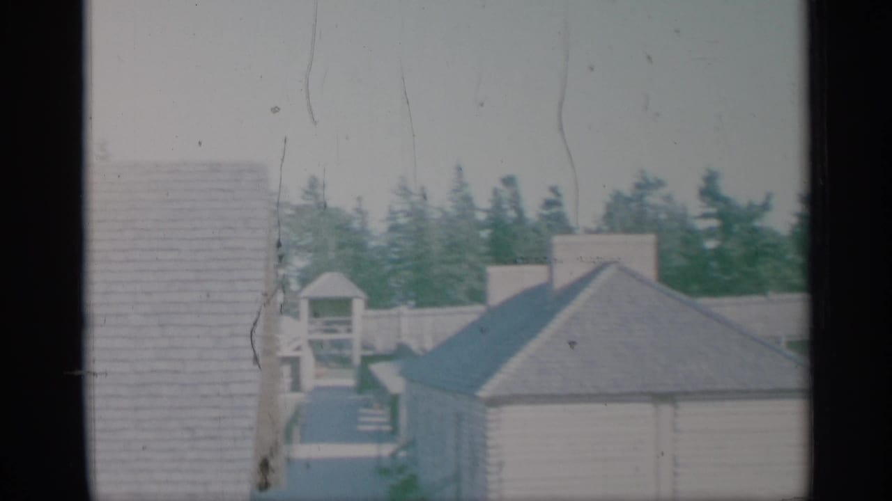 1959 New York 8mm frame showing a close wall, small roofed structure, house roof, evergreen trees, and faded film texture.