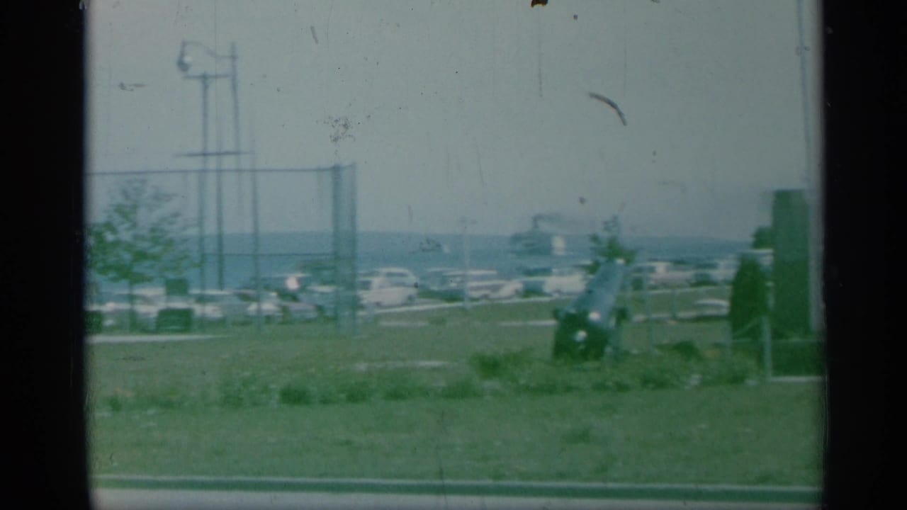 1959 New York 8mm view of grassy headstones, a chain-link fence, parked cars, a water tower, and distant water.