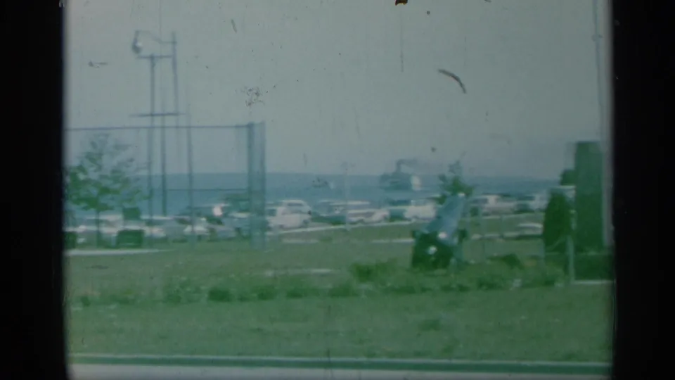 1959 New York 8mm view of grassy headstones, a chain-link fence, parked cars, a water tower, and distant water.