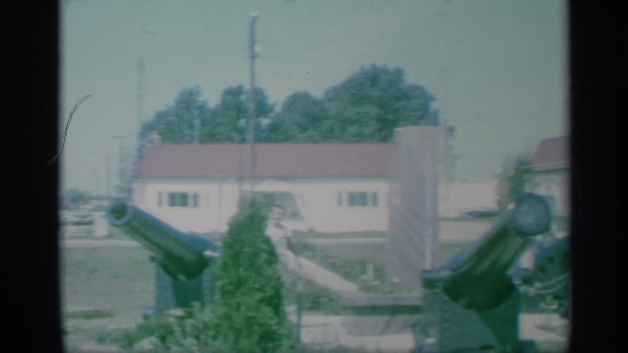 Cannons in a grassy military area with low buildings and a person partly visible nearby.