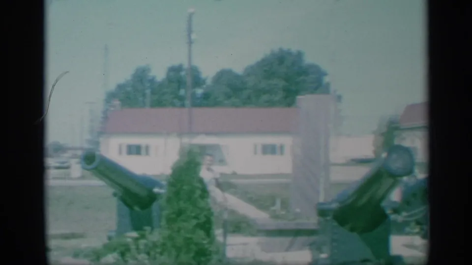 1959 New York 8mm frame showing cannons in a grassy military area, with low buildings, trees, and a person partly visible near the equipment.