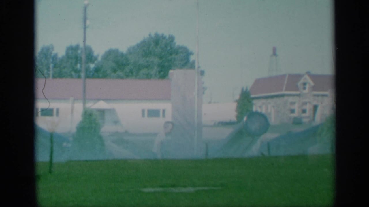 1959 New York 8mm frame with a cannon in a grassy military area, low buildings with red roofs, trees, and a pale sky in the background.