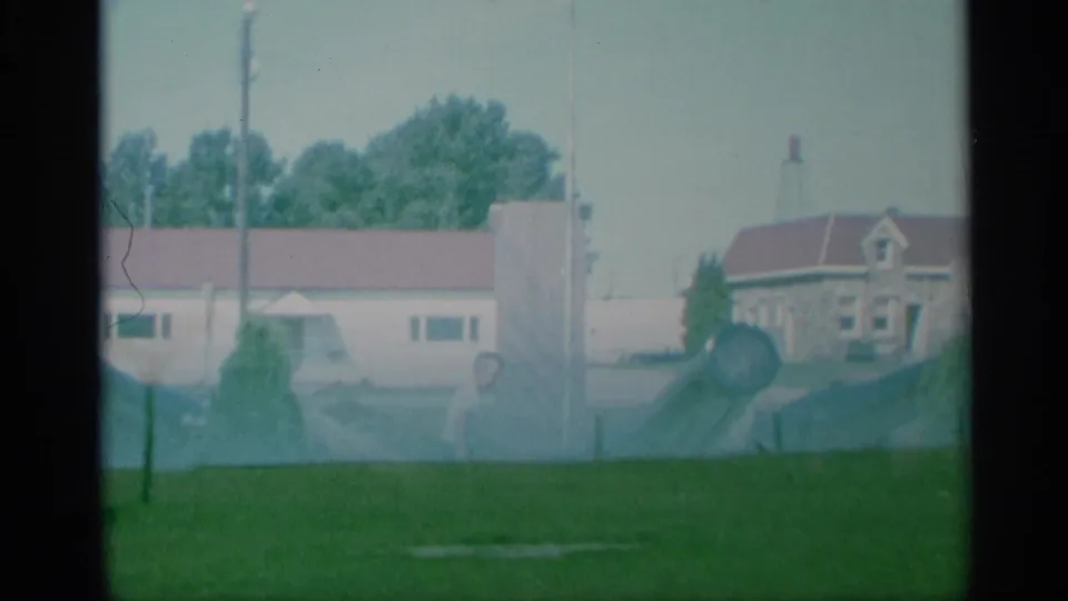1959 New York 8mm frame with a cannon in a grassy military area, low buildings with red roofs, trees, and a pale sky in the background.