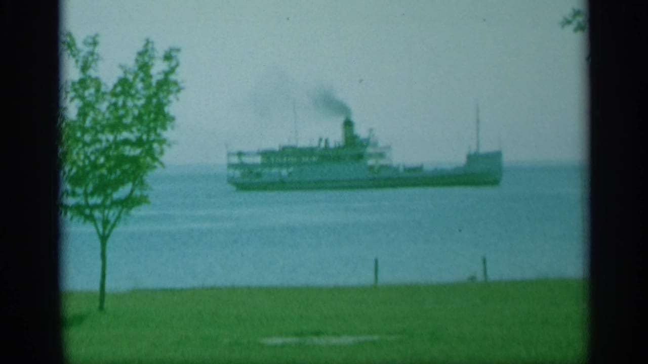1959 New York 8mm frame showing a steamship moving across open water beyond a small tree and grassy shoreline, with smoke rising from its stack.