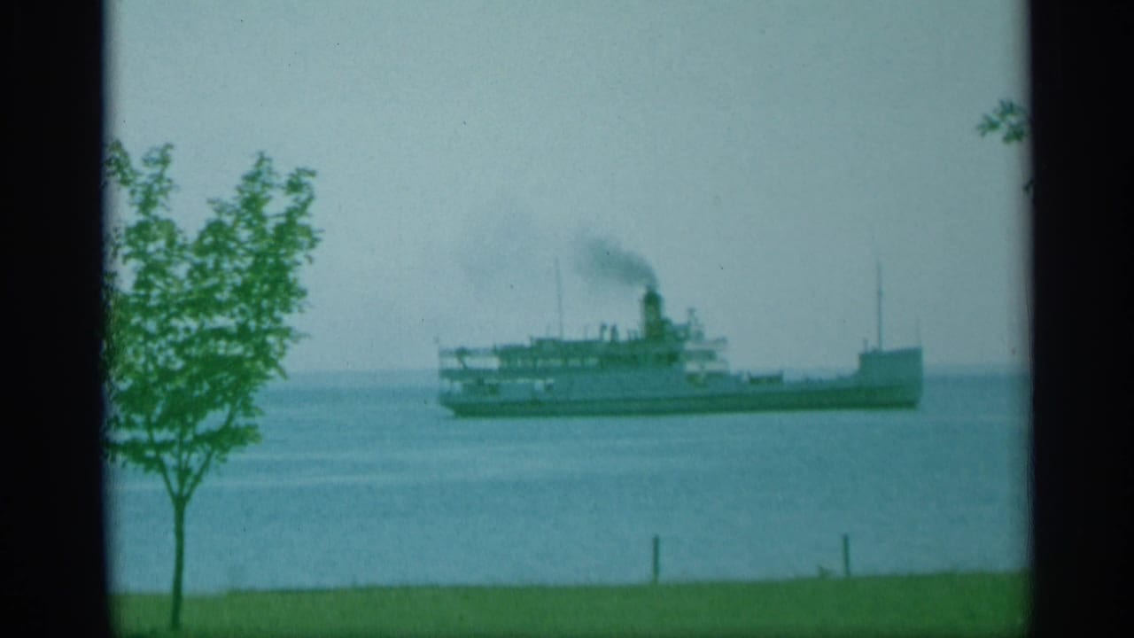 1959 New York 8mm frame of a steamship passing across open water, viewed from a grassy shoreline with a small foreground tree and soft faded color.