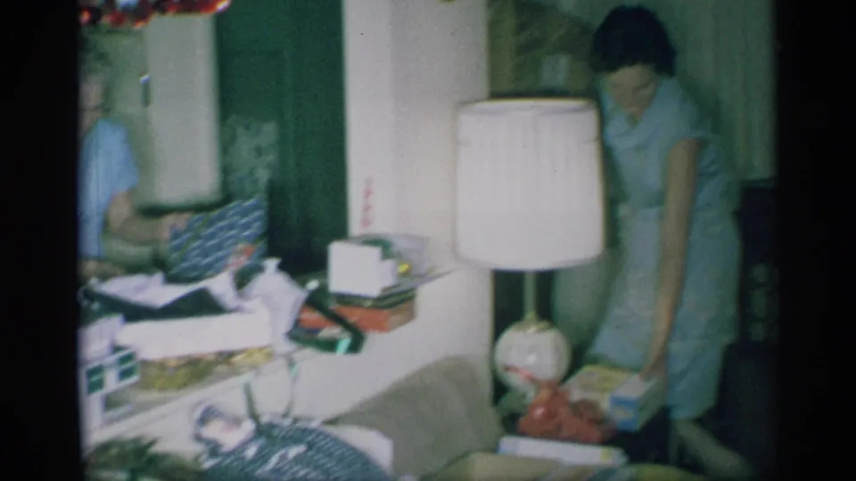 In a 1959 New York home-movie frame, a woman in a light blue dress handles presents beside a white lamp and cluttered furniture.
