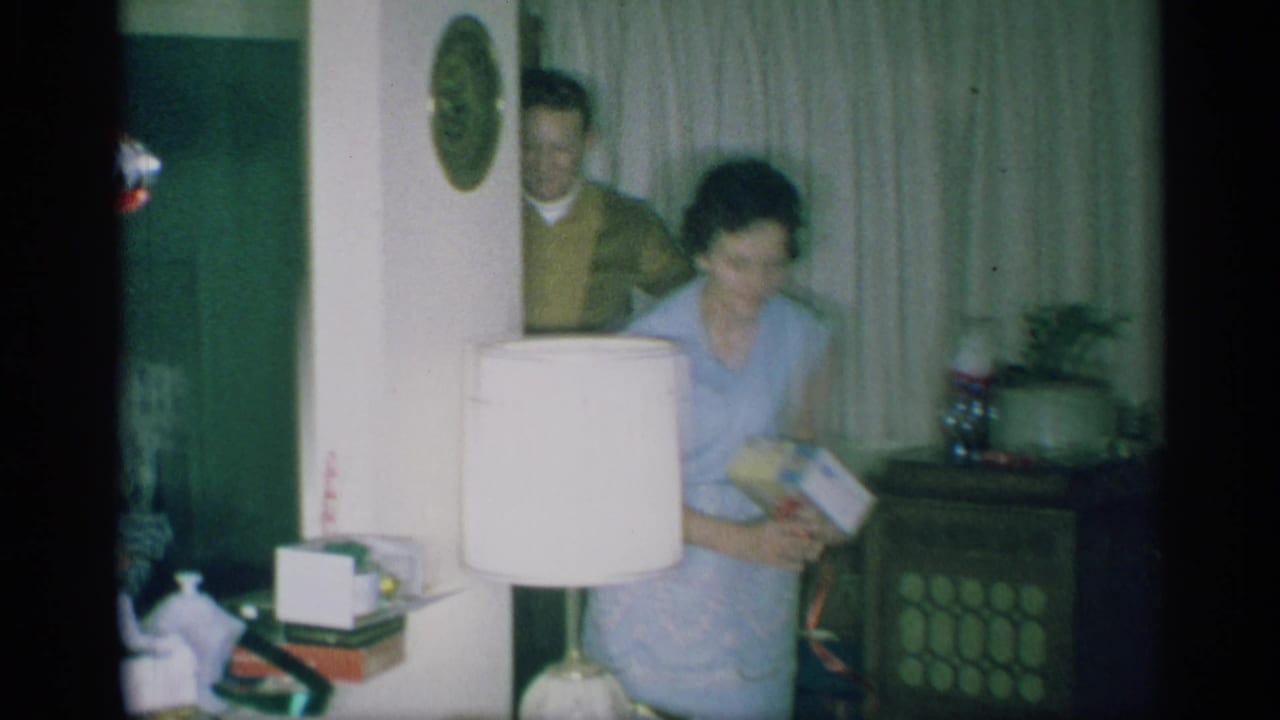 A woman in a light blue dress holds a present in a New York living room as a man in a brown sweater appears in the doorway.