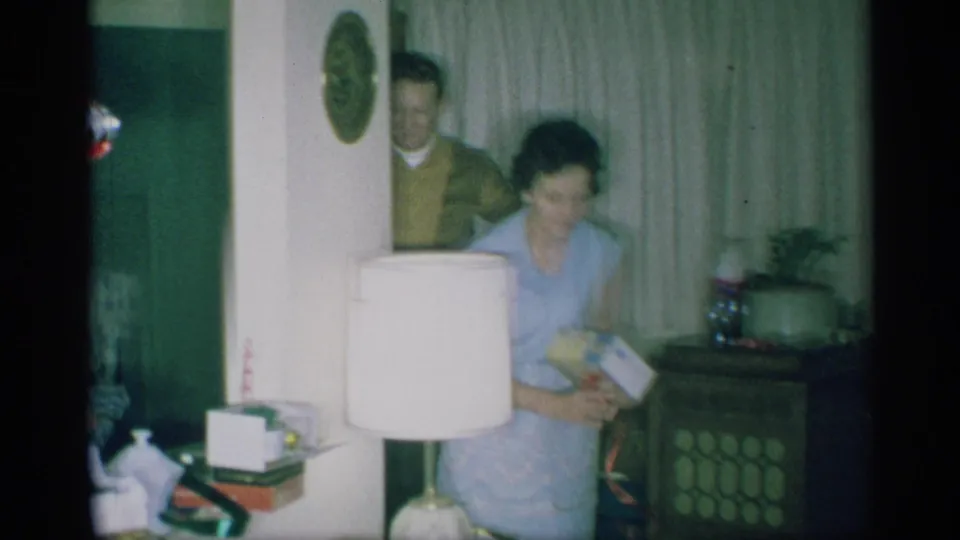 A woman in a light blue dress holds a present in a New York living room as a man in a brown sweater appears in the doorway.