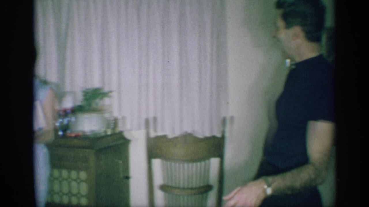 A man in a dark shirt stands near curtains, a chair, and a side table in the same 1959 New York Christmas living room.