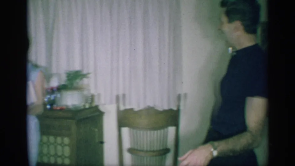 A man in a dark shirt stands near curtains, a chair, and a side table in the same 1959 New York Christmas living room.