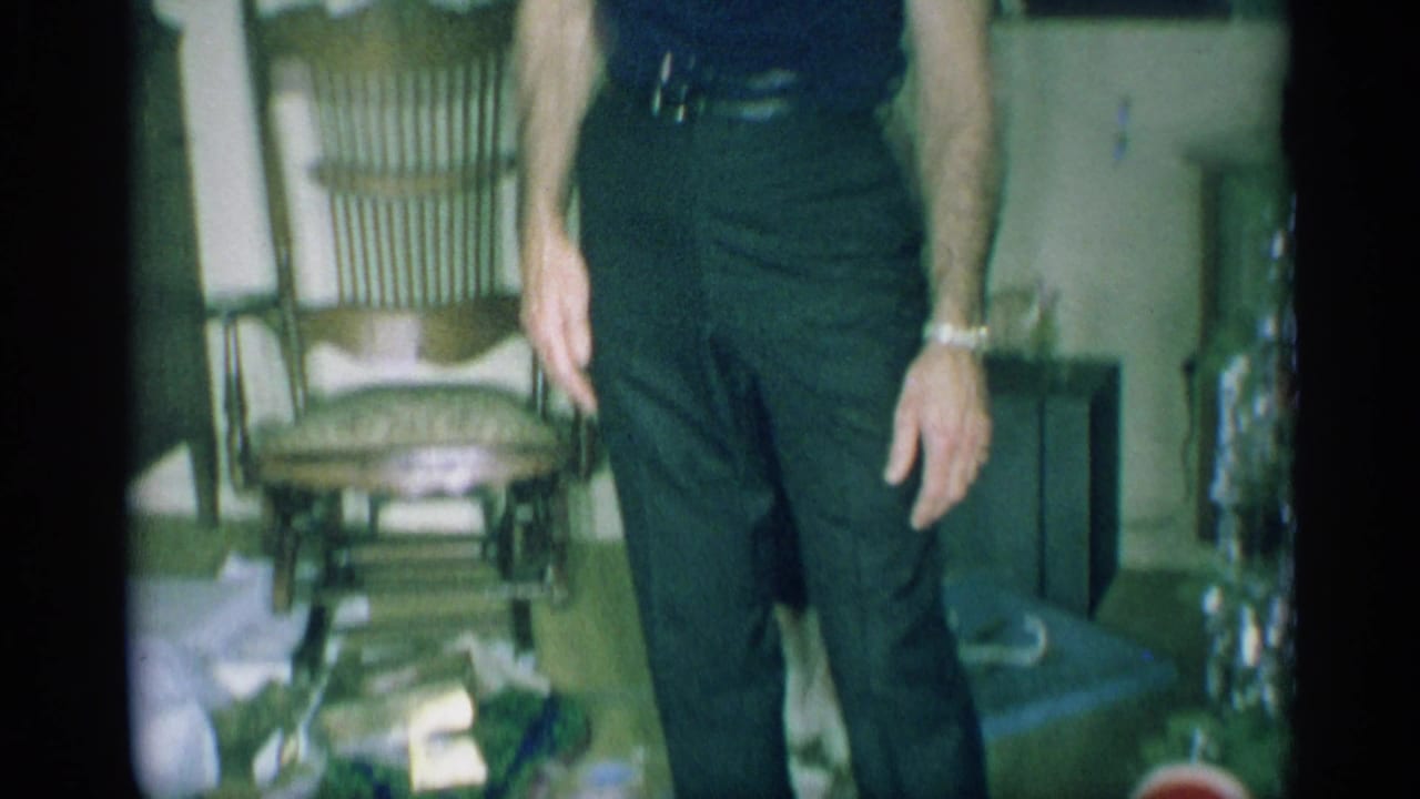 A man in dark clothing is partly framed near mid-century furniture, loose paper, and a small Christmas tree or holiday decoration.