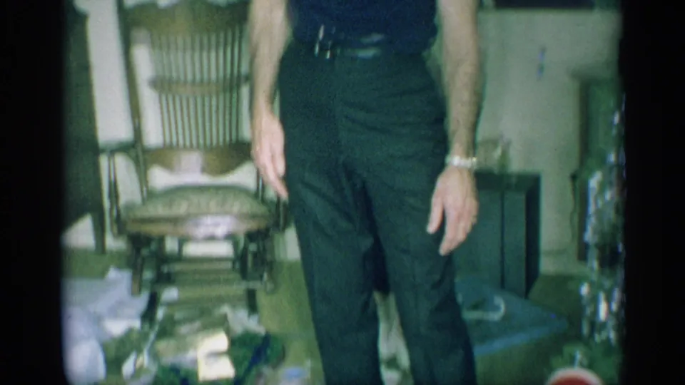 A man in dark clothing is partly framed near mid-century furniture, loose paper, and a small Christmas tree or holiday decoration.