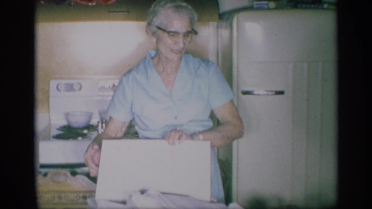 1959 New York kitchen scene with an elderly woman in glasses and a light blue dress holding a white box near a retro stove and refrigerator.