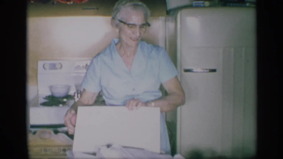 1959 New York kitchen scene with an elderly woman in glasses and a light blue dress holding a white box near a retro stove and refrigerator.