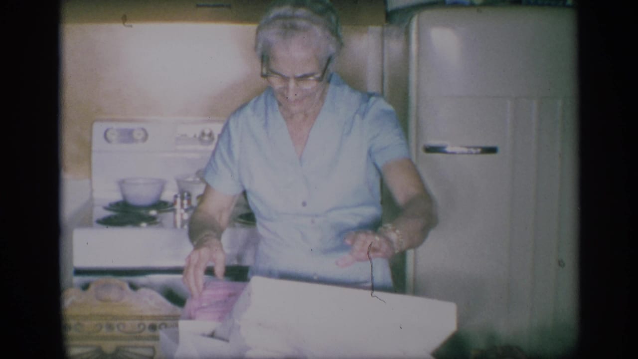 The woman looks down into a white box in the 1959 New York kitchen, with a stove, refrigerator, and household items behind her.