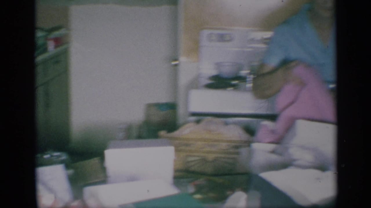 A cropped 1959 New York kitchen frame shows the woman moving beside a table with a white box, fabric, and scattered household items.