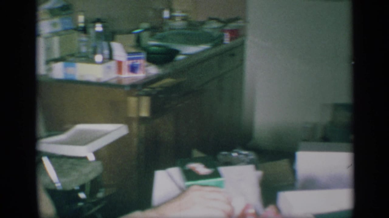 1959 New York home-movie frame showing a partially framed seated man in a kitchen with household boxes and a small tray chair nearby.