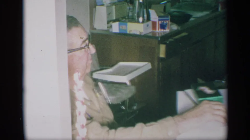 1959 New York home-movie frame of an older man with glasses seated in a kitchen near counters, boxes, and a small tray chair.