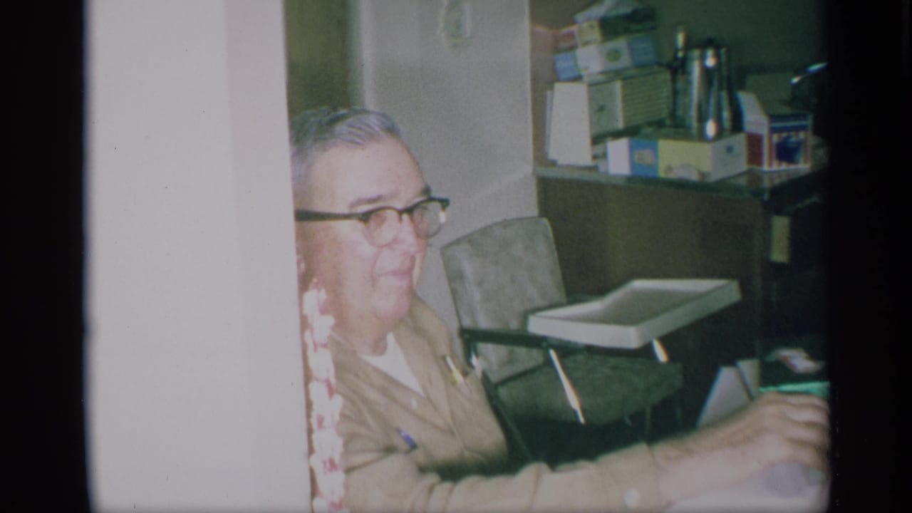 1959 New York home-movie frame showing an older man with glasses seated in a kitchen, partly obscured by a light wall or doorway edge.