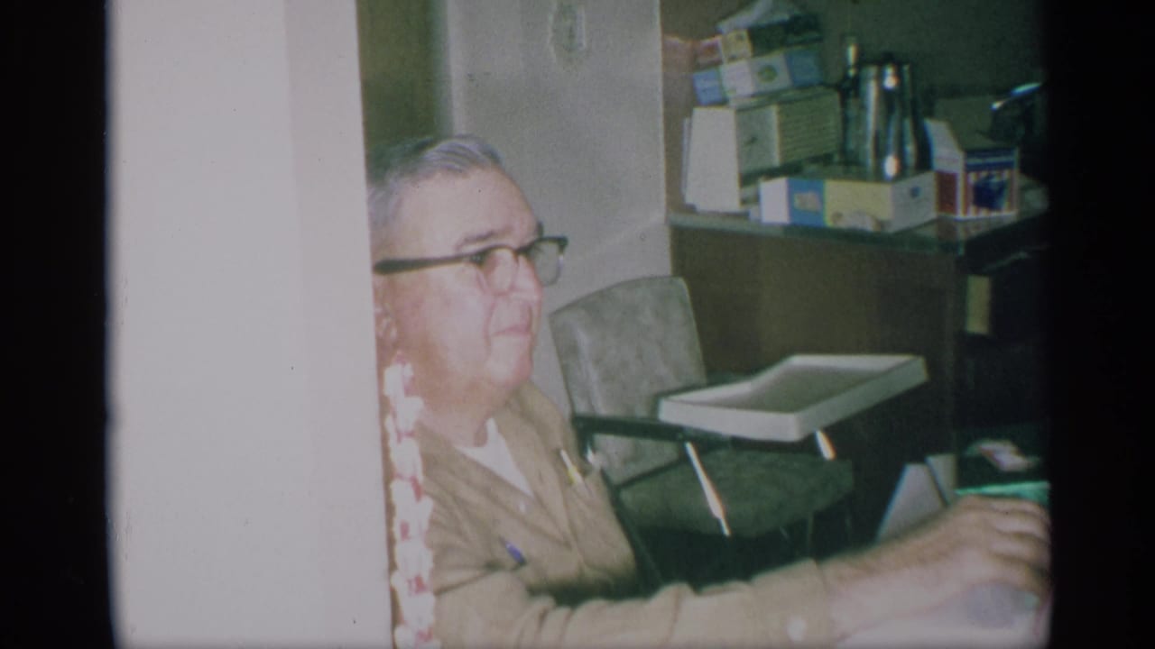 1959 New York home-movie frame of an older man in glasses seated in a private kitchen with boxed goods and a small tray chair behind him.