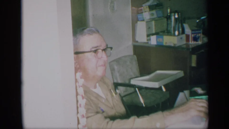 1959 New York home-movie frame of an older man in glasses seated in a private kitchen with boxed goods and a small tray chair behind him.