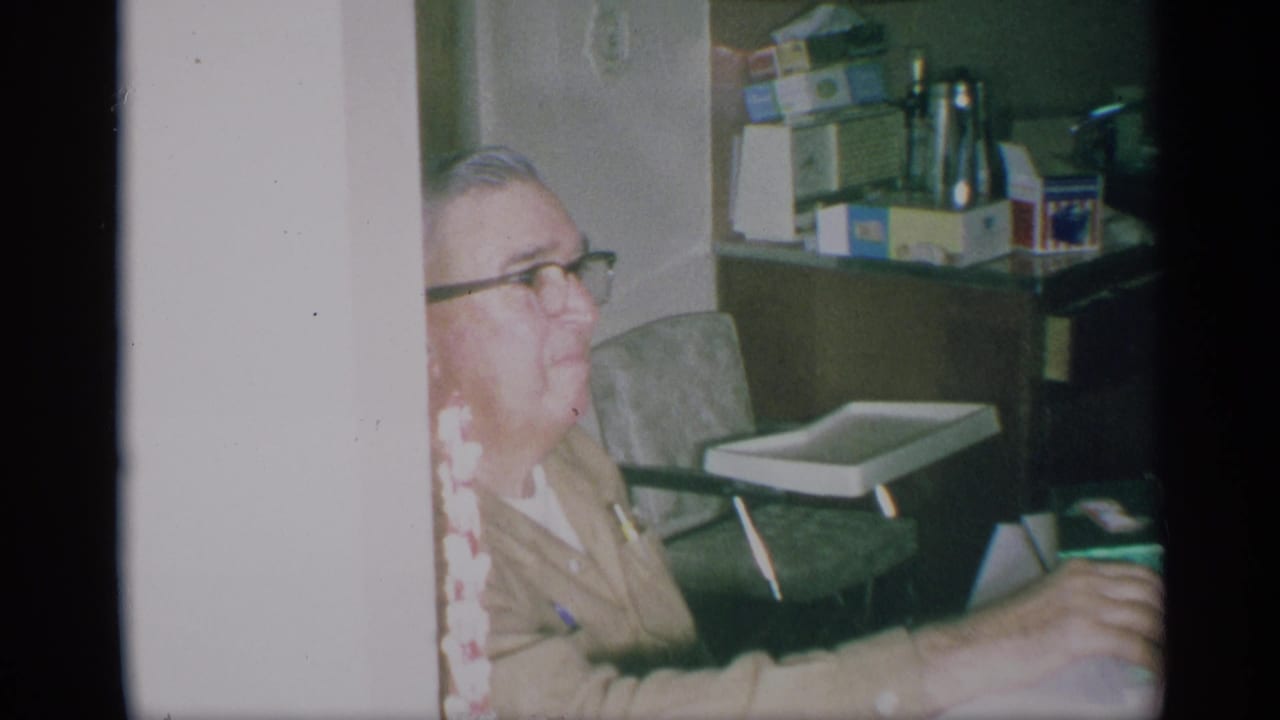 In a 1959 New York home-movie frame, an older man with glasses sits in a private kitchen near shelves, a chair, and a small tray.