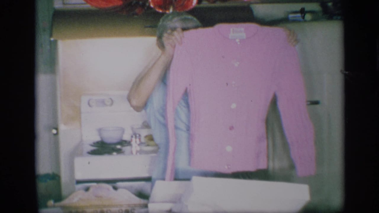 Older woman holds up a pink cardigan in a kitchen with a stove and gift box nearby.