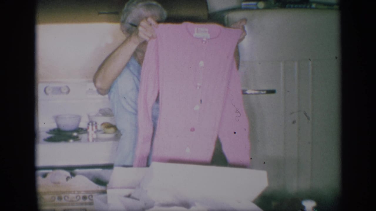 The woman displays the pink cardigan toward the camera in the home kitchen, with the stove, refrigerator, and open box behind it.