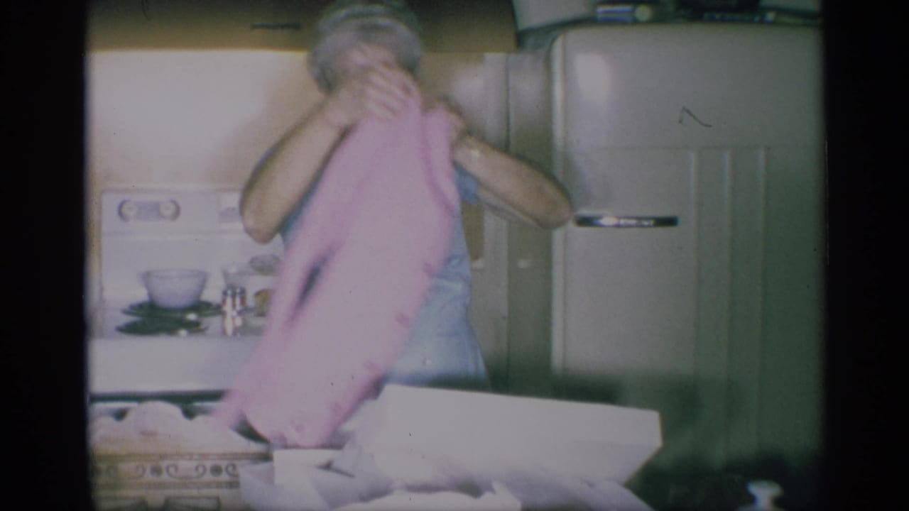 The woman lowers or folds the pink cardigan beside the open gift box in the 1959 New York kitchen, with a refrigerator behind her.