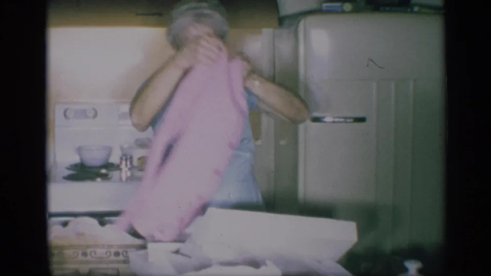 The woman lowers or folds the pink cardigan beside the open gift box in the 1959 New York kitchen, with a refrigerator behind her.