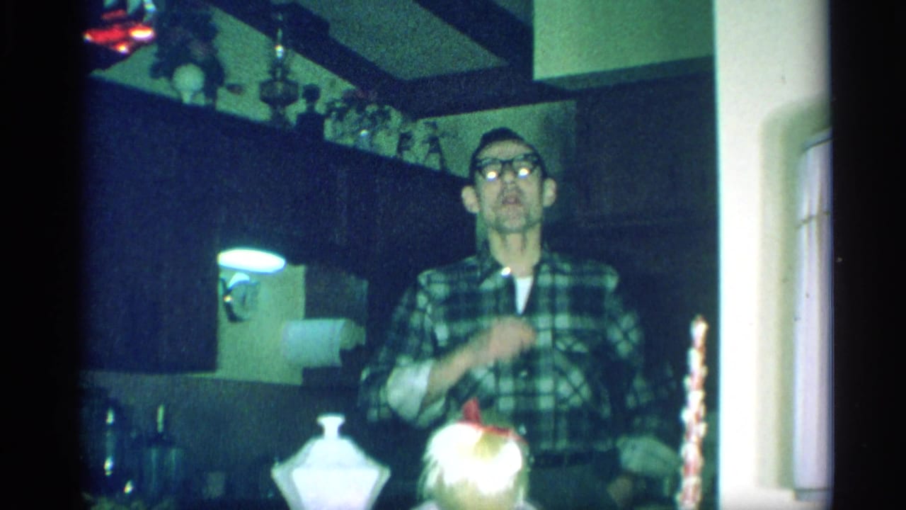 A man in glasses and a plaid shirt stands in a dim 1959 New York kitchen with cabinets and decorative items behind him.
