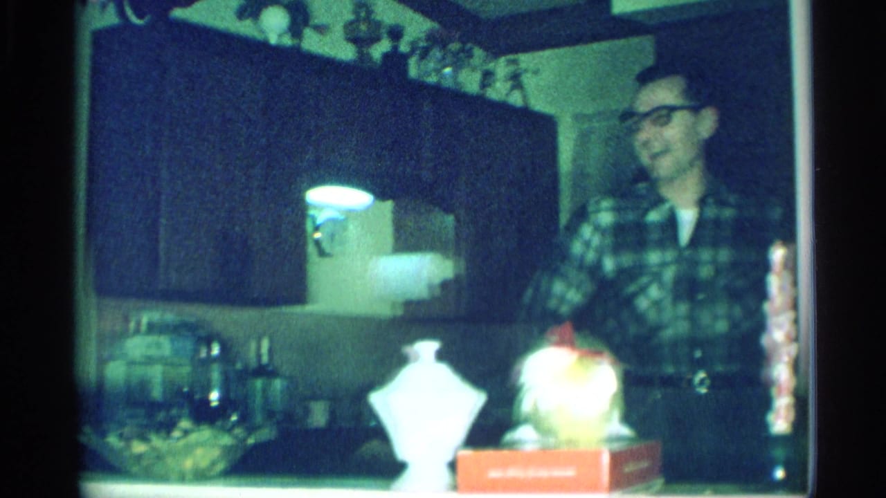 The 1959 New York home-movie frame shows a smiling man in a plaid shirt beside kitchen cabinets and tabletop ornaments.