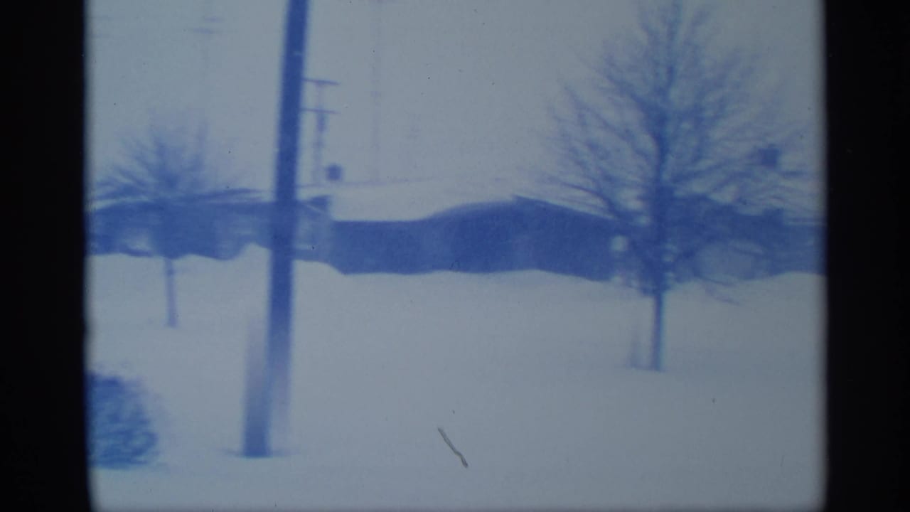 Blue-tinted 8mm frame from New York showing utility poles, bare trees, and snow around low suburban buildings.