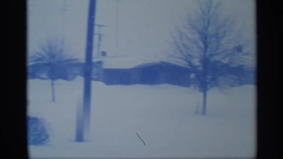Blue-tinted 8mm frame from New York showing utility poles, bare trees, and snow around low suburban buildings.