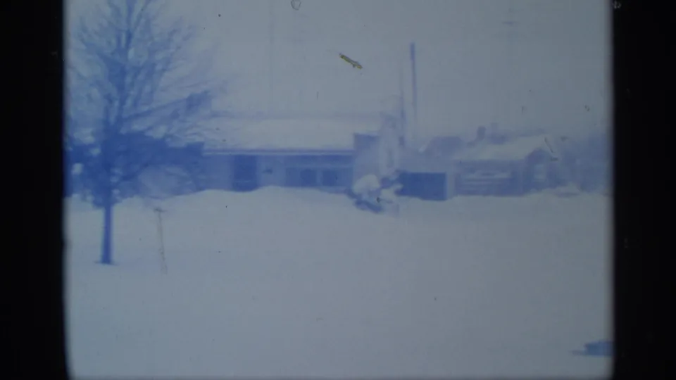 1959 New York 8mm frame showing a snow-covered rural yard, bare trees, and distant low buildings with a blue film cast.