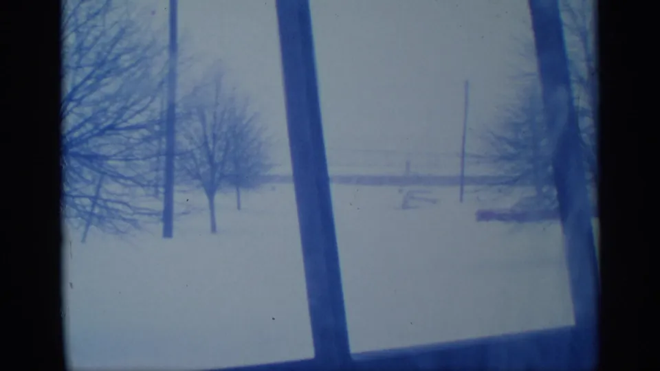 1959 New York 8mm frame showing a snow-covered rural view with bare trees, utility poles, and faint structures beyond the field.