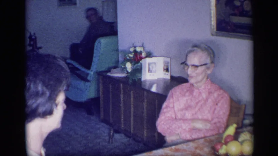 1959 New York interior home-movie frame with two women seated in a living room, one in glasses and a pink blouse near a table.