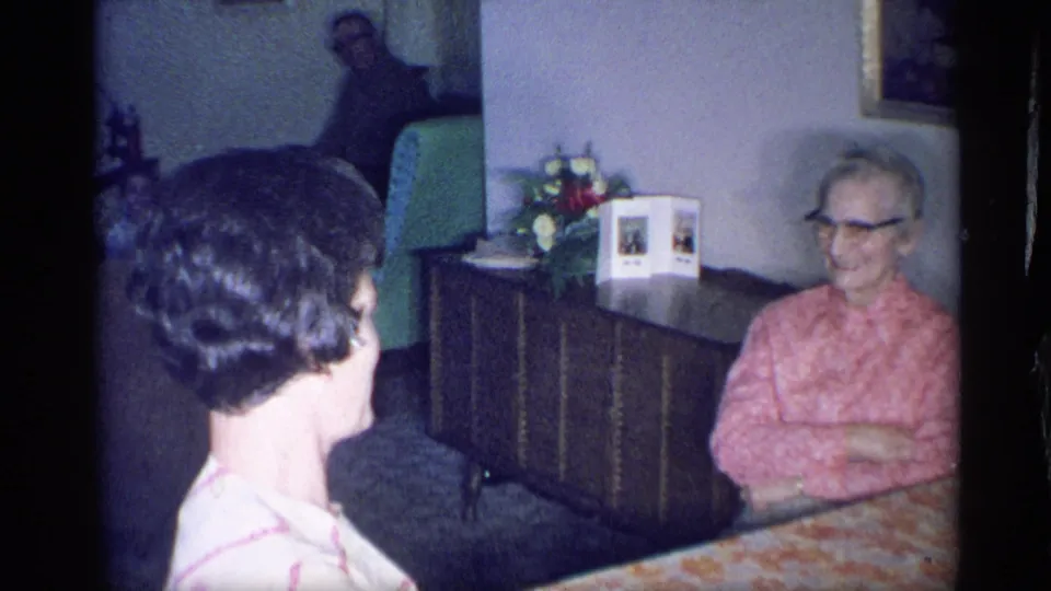 1959 New York home-movie frame with a younger woman seen from behind while an elderly woman in a pink blouse smiles at the table.