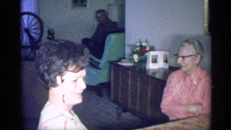 1959 New York home-movie frame showing a younger woman smiling in profile, with an elderly woman seated near a dark wood cabinet in the living room.