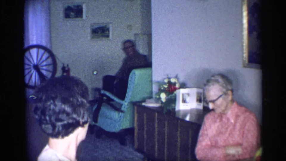 1959 New York home-movie frame showing two women near a table, a man in a green chair, framed pictures, flowers and a spinning wheel in a private interior.