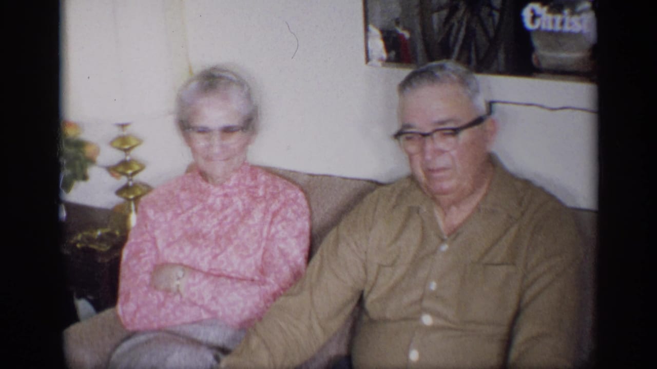 Older woman and man with glasses sit together on a living room couch.