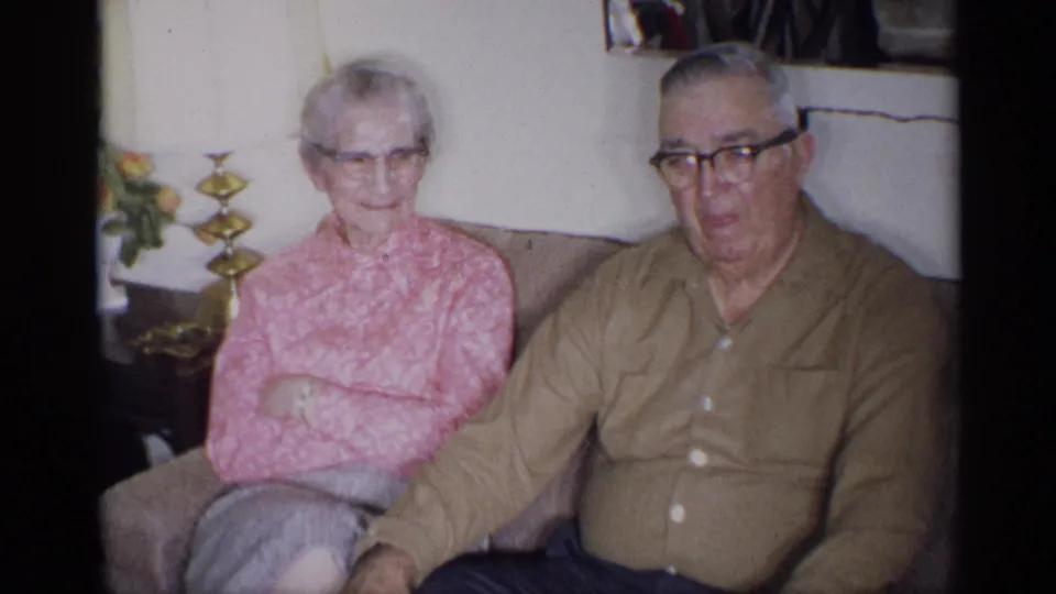 The woman appears to smile faintly as she sits beside the man on the couch in the private home interior.