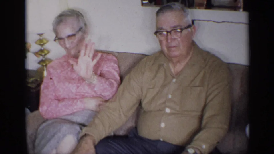 The older woman raises her hand toward the lens while the man sits beside her, both visible in the 1959 New York home interior.