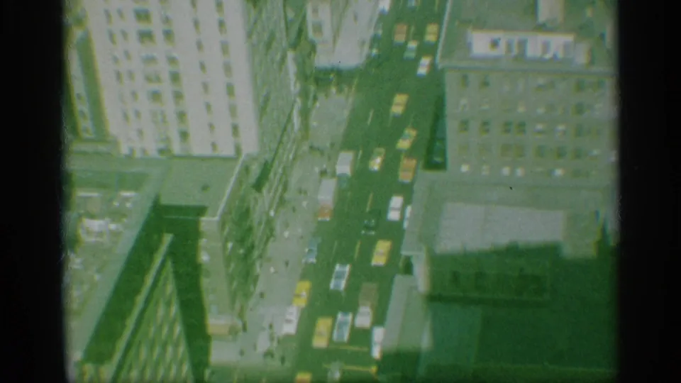 1959 New York high-angle 8mm view looking down on tall buildings, a straight street, parked or moving cars, rooftops, and deep shadows.